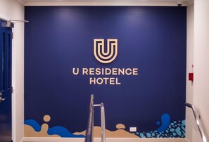 Una pared con el logotipo y el nombre del Hotel U Residence, un pasamanos de metal y una puerta.