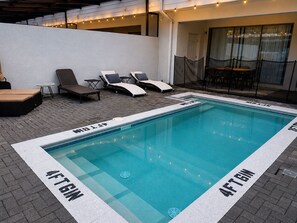 A swimming pool with lounge chairs and a table, surrounded by a brick patio.
