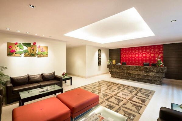 A modern living room with a sofa, ottoman, and coffee table, a reception desk with a red patterned wall, and a skylight. - The Lotus Service Apartments (Chennai)