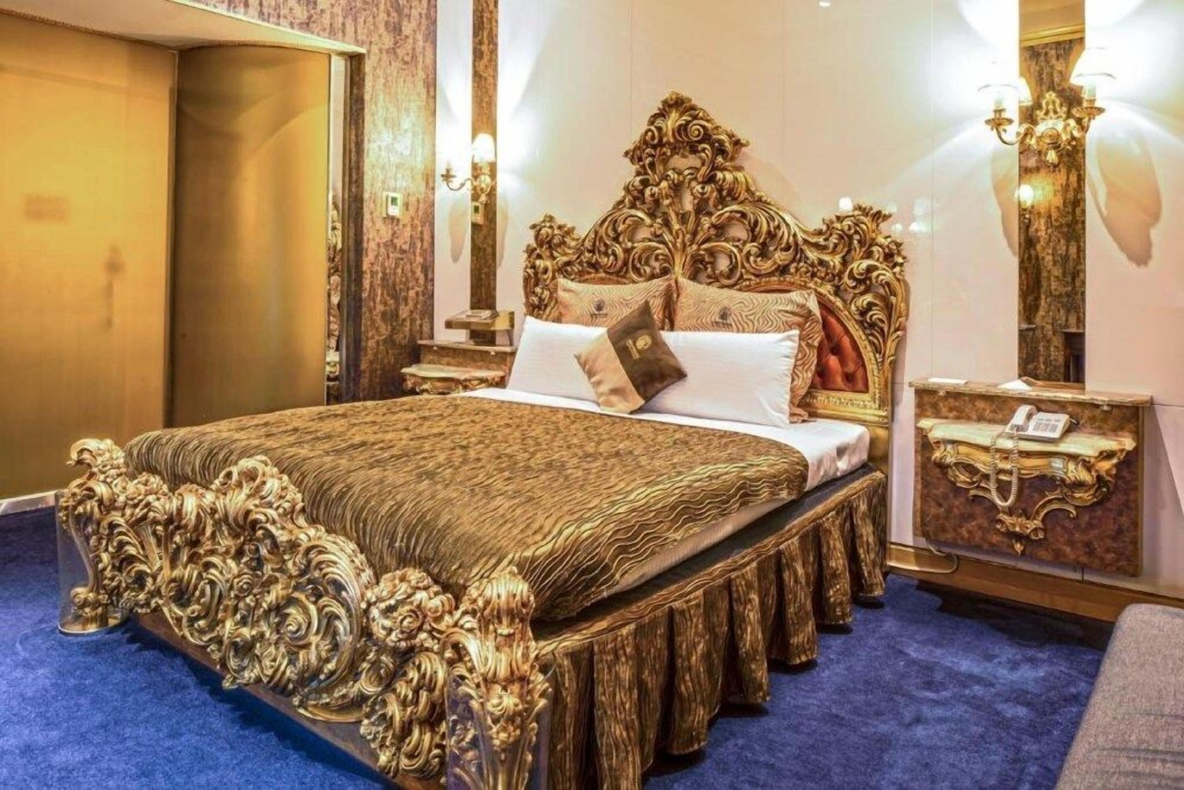 A luxurious bed with ornate headboard and footboard, a bedside table with a telephone, and a wall-mounted lamp.