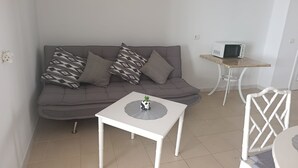 En moderne stue med en grå sofa, mønstrede puder, et hvidt sofabord med en panda-figur, et hvidt sengetæppe med en mikroovn, og en hvid stol med en vævet ryg.