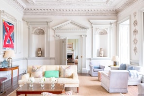 A spacious living room with a high ceiling, ornate moldings, and a grand staircase leading to another room.