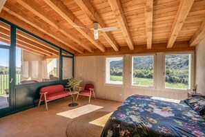 Ein Schlafzimmer mit Holzboden, einem Bett mit floralem Überwurf, einem roten Sofa, einem kleinen Tisch mit einer Vase und großen Fenstern mit Blick auf Grünflächen.