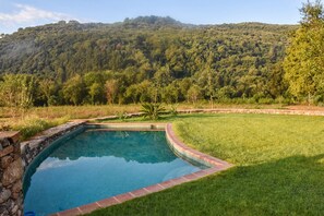 Une piscine bordée de pierres, entourée d’une pelouse bien entretenue et offrant une vue sur une colline boisée.