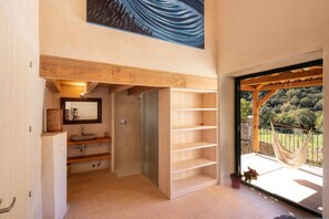 Une salle de bain moderne avec un meuble-lavabo en bois, un miroir et une douche. Une grande fenêtre offre une vue sur les arbres, et un hamac est installé à l’extérieur.