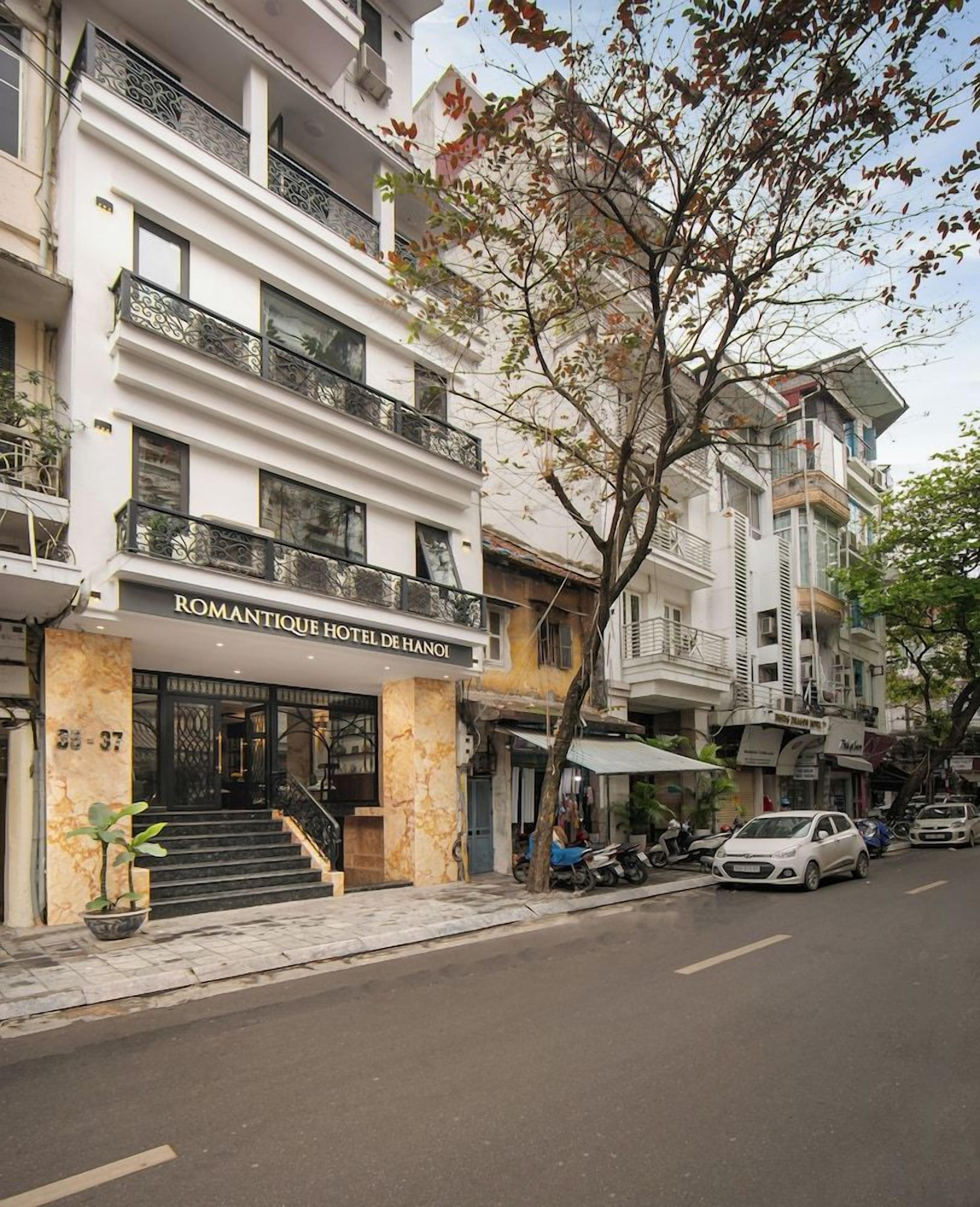 A multi-story hotel with a balcony on the second floor, a staircase leading to the entrance, and parked cars along the street.