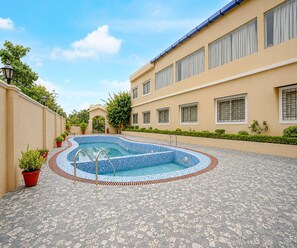 Una piscina con bordeado de azulejos, rodeada por un edificio con múltiples ventanas y una entrada protegida por un portón.