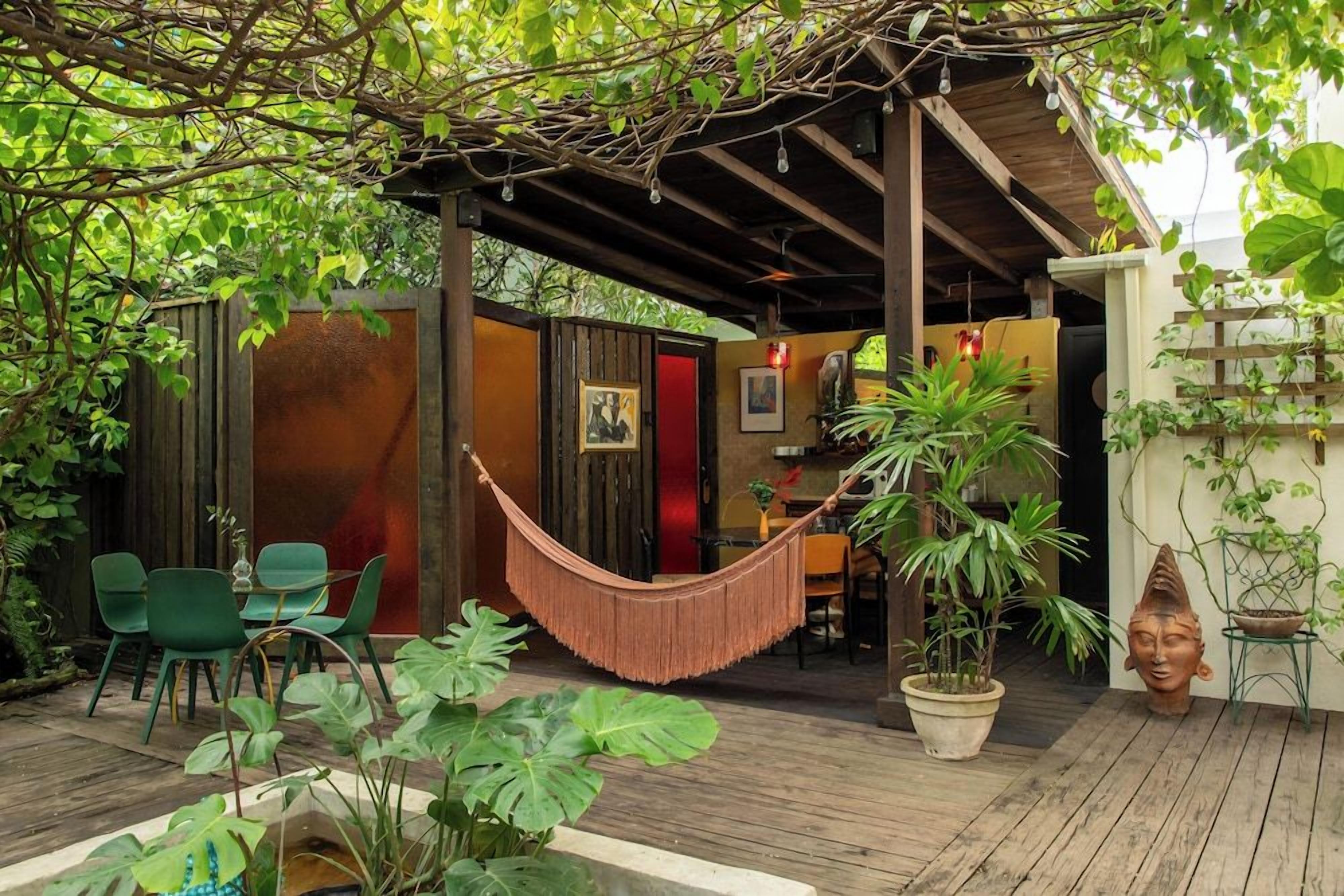 A covered outdoor seating area with wooden flooring, green chairs, a hammock, and potted plants.