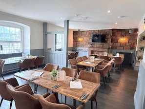 A restaurant with multiple tables and chairs, a brick wall with a mounted TV, and a window providing a view. - The Miners Rest at Radstock Hotel (Radstock)