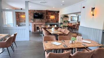 Un restaurante con mesas y sillas de madera, una pared de ladrillo con chimenea y un televisor montado en la pared.