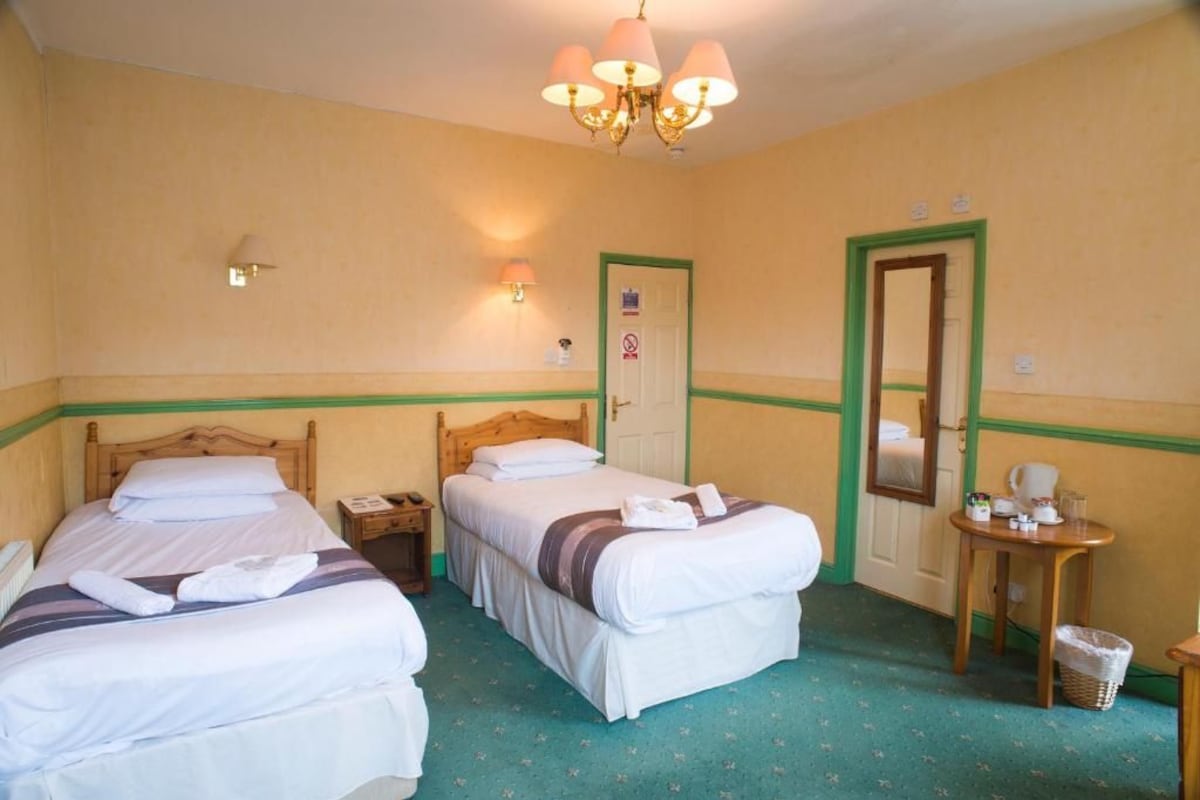 A hotel room with two beds, a wooden headboard, a small table with a chair, a mirror, and a chandelier.