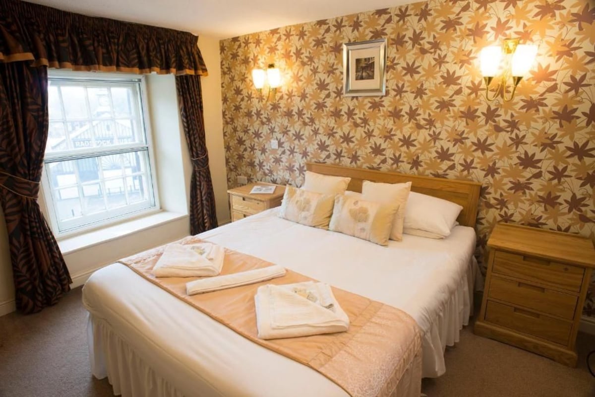 A hotel room with a large bed, two bedside tables, a window with curtains, a wall with a leaf pattern, and two wall-mounted lights.