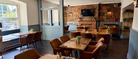 A restaurant with wooden tables and chairs, a brick wall with a television, and a fireplace.