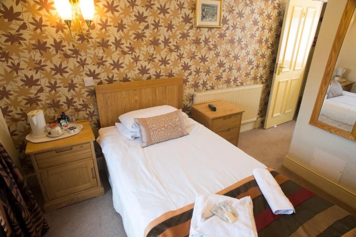 A hotel room with a bed, bedside tables, a nightstand, a radiator, a mirror, and a patterned wallpaper.