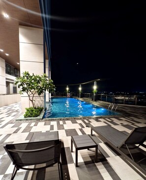 Un área de piscina en la azotea con sillones de descanso y vistas a la ciudad de noche.