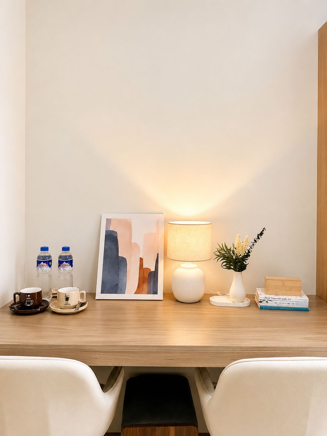 A hotel desk with a framed abstract painting, two water bottles, a coffee cup, a white lamp, a small vase with flowers, and a stack of books.