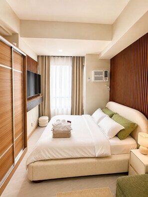 A hotel room with a large bed, a flat-screen TV, an air conditioning unit, and a bedside table with a lamp. - Cozy studio in lovely Quezon City with refreshing AC (Quezon City)