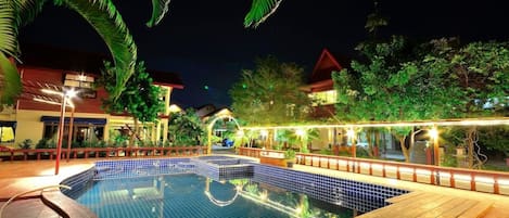 A swimming pool with a tiled deck, surrounded by greenery and buildings.