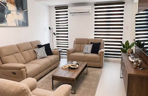 Una sala de estar con un sofá color beige, un sofá más pequeño, una mesa de centro y una mesita auxiliar con objetos decorativos.