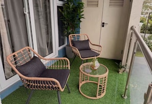 Un balcón con dos sillas de mimbre, una pequeña mesa redonda, una planta en maceta y vistas de un edificio con una puerta y ventanas.