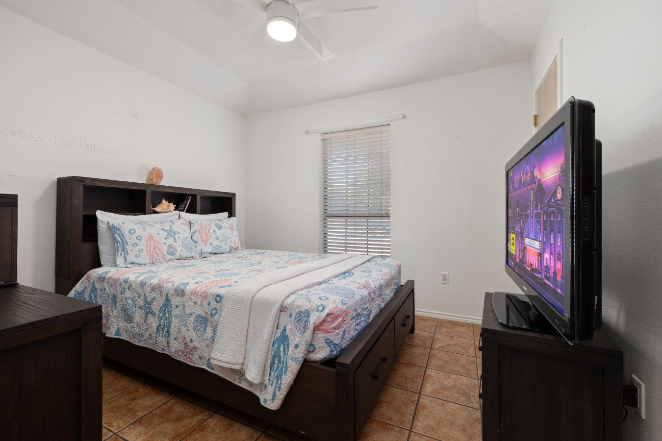 A bedroom with a bed, a television, a window with blinds, and a ceiling fan.