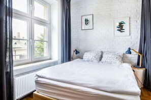 A bedroom with a large bed, a white brick wall, two framed artworks, and a window with curtains.