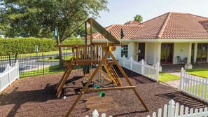 A playground with a swing set and a fenced area, a building with a tiled roof, and a white picket fence. - 5BR Tuscan Hills Villa Pool & Themed Rooms (Davenport)