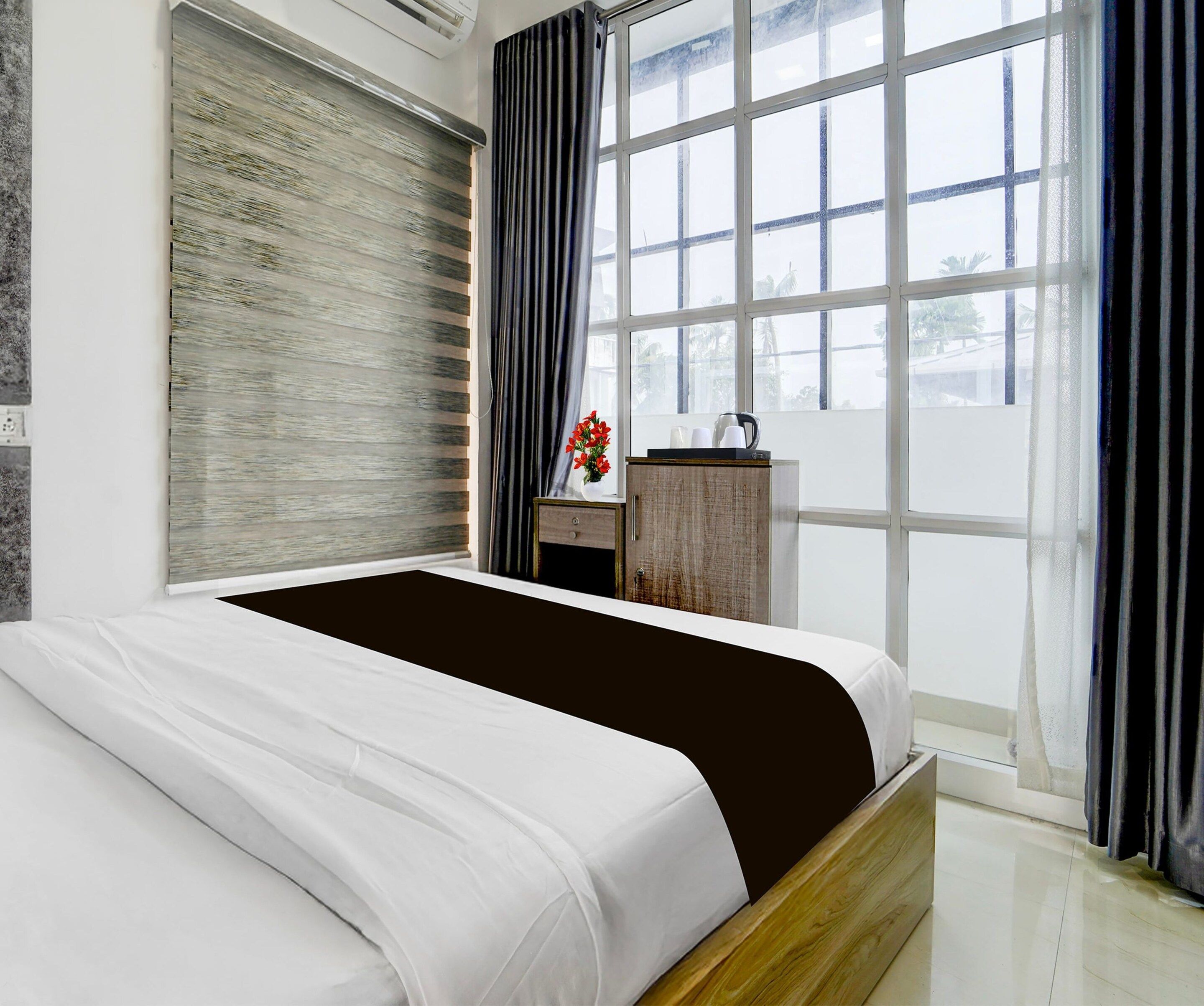 A modern bedroom with a large bed, a wooden nightstand, a small table with a vase of flowers, and a view of the outdoors through a large window.