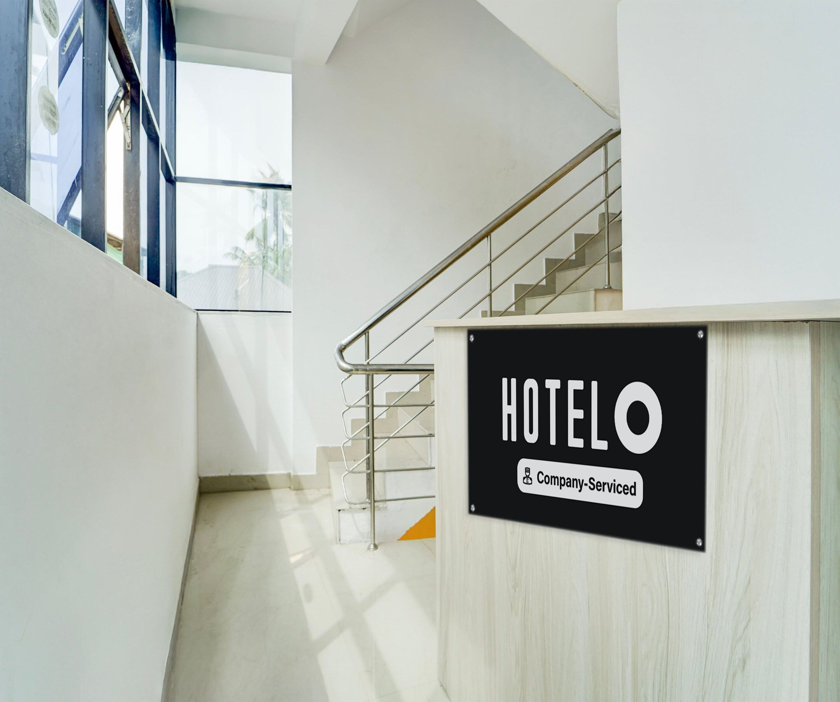 A hotel reception area with a staircase leading to upper floors, a sign indicating 'HOTEL & Company-Serviced', and a view of the outside through a glass door.
