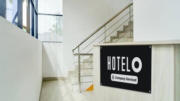 A hotel reception area with a staircase leading to upper floors, a sign indicating 'HOTEL & Company-Serviced', and a view of the outside through a glass door.
