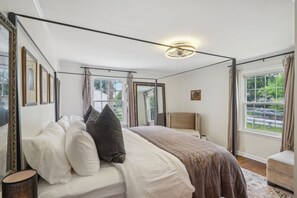 A bedroom with a bed, a chair, a bedside table, a mirror, and a window with curtains. - Broad Ripple Retreat | Stylish 3BR + Walkable (Indianapolis)