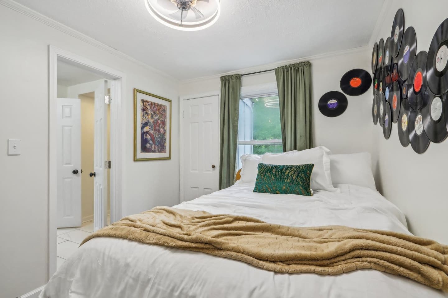 A bedroom with a bed, a wall of vinyl records, a door, and a window with curtains.