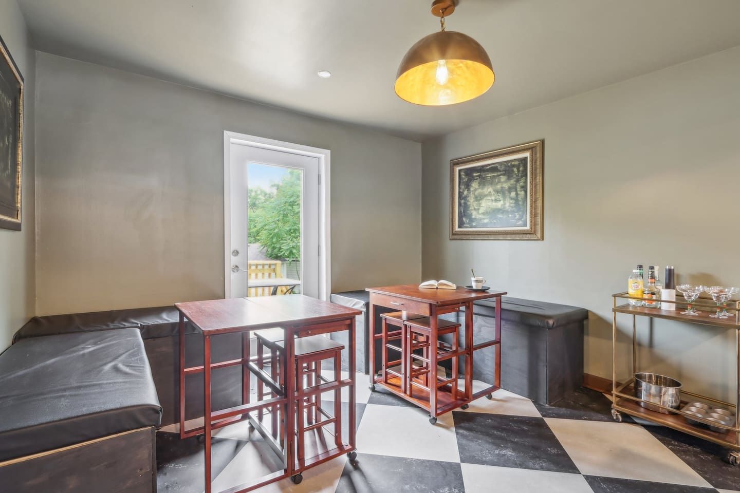 A room with a dining table, chairs, a bench, a small table with a book and a cup, a shelf with bottles and glasses, and a door leading to a balcony.