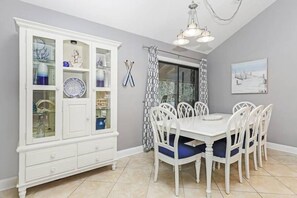 Un coin repas avec une table et des chaises blanches, un meuble avec des portes en verre et une fenêtre agrémentée de rideaux à motifs.