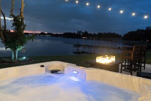 Uma banheira de hidromassagem com lareira, cercada por cadeiras e uma mesa, com vista para um lago calmo com um píer e árvores ao fundo.