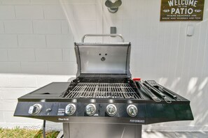 Una parrilla Char-Broil Performance con cuatro quemadores, una tapa y un asa, frente a una pared blanca con un cartel que dice 'Bienvenidos al Patio'.