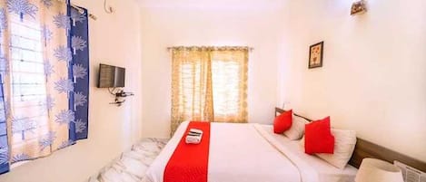 A hotel room with a bed, a bedside table, a lamp, a window with curtains, and a wall-mounted TV.