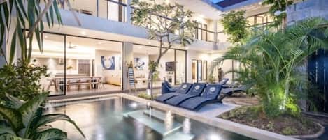 A modern two-story building with a swimming pool, outdoor seating, and lush greenery.