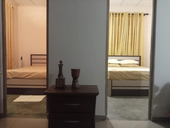 A room with two beds, a wooden nightstand with a statue and a cup, and a door leading to another room.