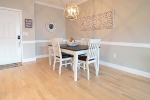 A dining area with a wooden table, white chairs, a decorative wall piece, and a wall clock. - Folly Road Retreat - Folly Beach Monthly Rental (Folly Beach)