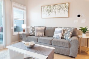 A living room with a grey sofa, patterned cushions, a small table with a bowl, a wall-mounted picture, and a potted plant. - Folly Road Retreat - Folly Beach Monthly Rental (Folly Beach)