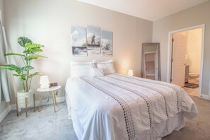 A bedroom with a bed, a bedside table with a lamp, a potted plant, and wall art. - Folly Road Retreat - Folly Beach Monthly Rental (Folly Beach)