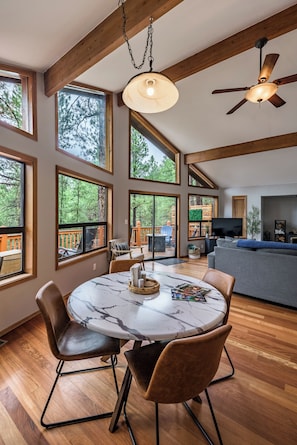 A spacious dining area with a round marble table, wooden flooring, and large windows offering a view of a deck and trees. - Stylish Zen Mountain Villa: Hot Tub, Games & Views (Flagstaff)