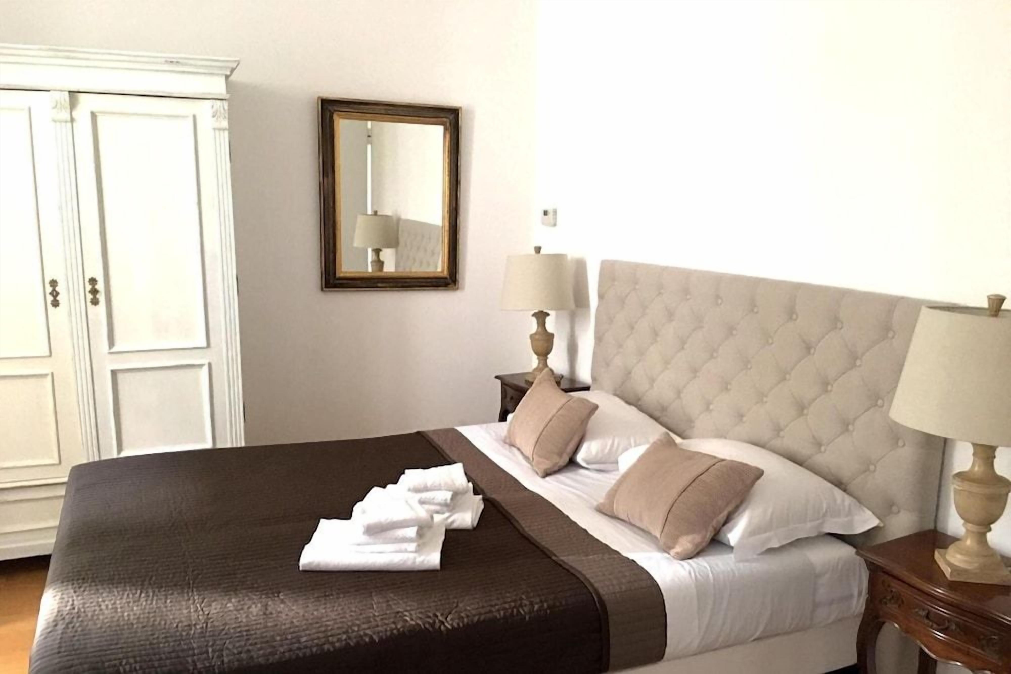 A hotel room with a bed, a nightstand, a lamp, a mirror, and a wardrobe.
