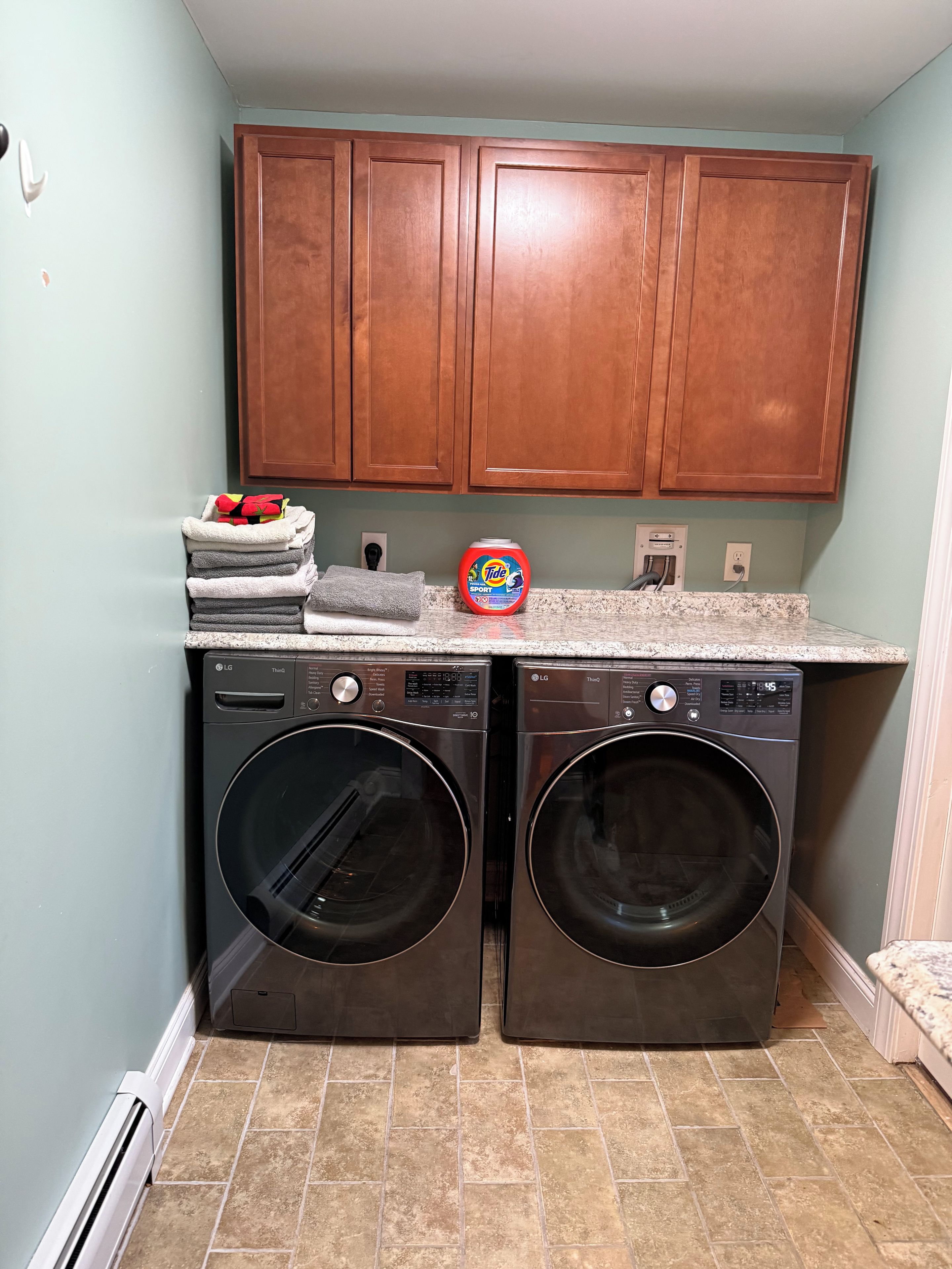 A laundry area with two front-loading LG washers, a stack of folded towels, and a Tide detergent bottle.