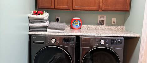 A laundry area with two front-loading LG washers, a stack of folded towels, and a Tide detergent bottle.