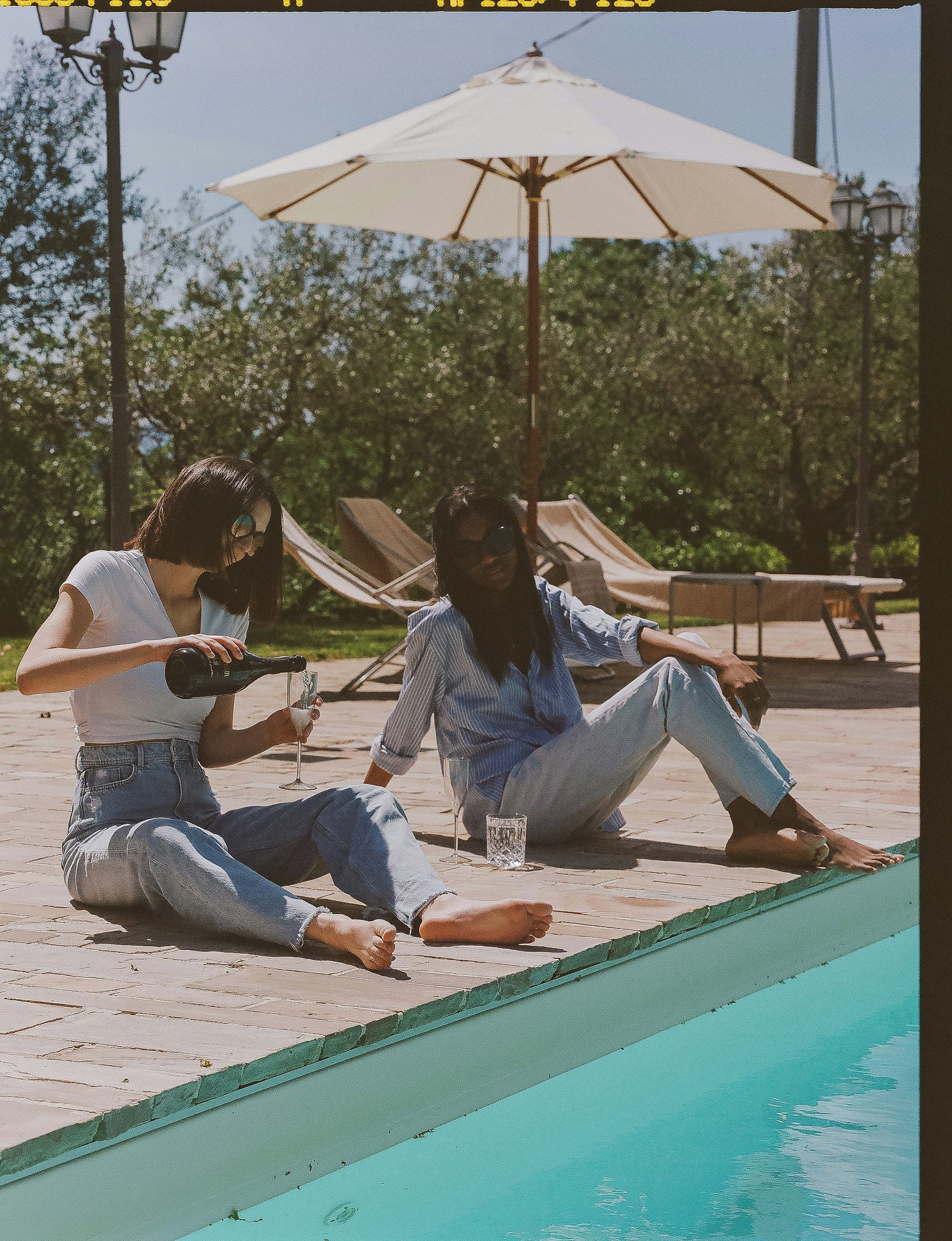 Two individuals sitting by a poolside with lounge chairs and an umbrella in the background.