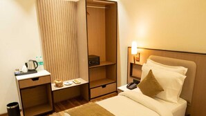 Quarto de hotel com cama, mesa de cabeceira, armário e pequena área de trabalho com chaleira e garrafas.