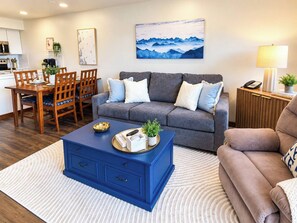 A living room with a blue coffee table, grey sofa, and a dining area with wooden chairs. - Highland House 317 Ski In Out Village Hot Tub Wifi (Snowshoe)