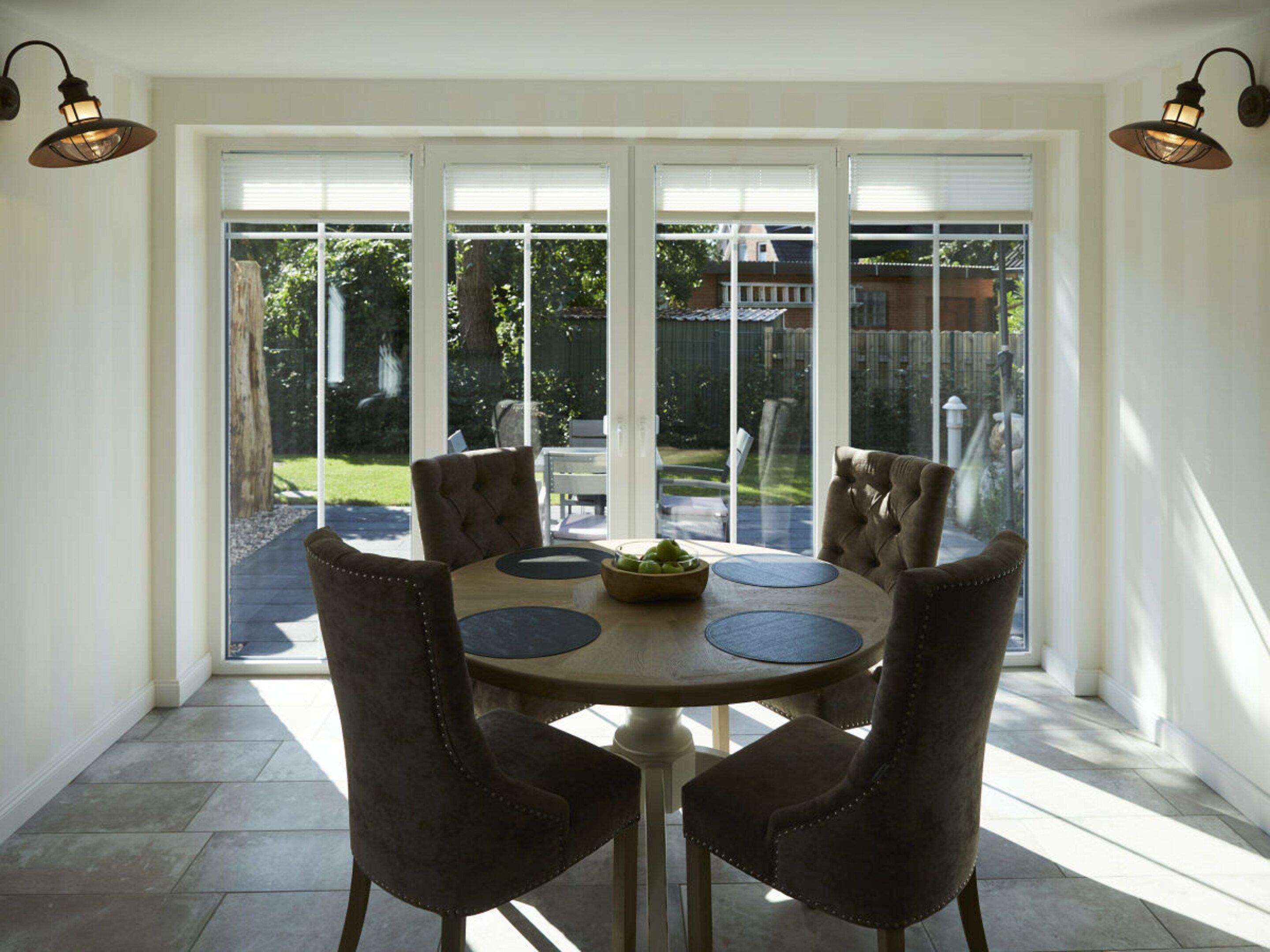 A dining area with a round table, six chairs, and a view of a backyard through large windows.